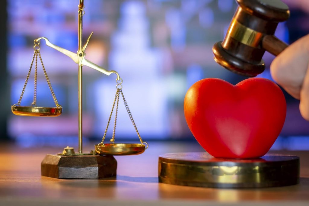 A red heart sits between a gavel and a sound block, next to a hanging scale that is tipped heavily to one side.