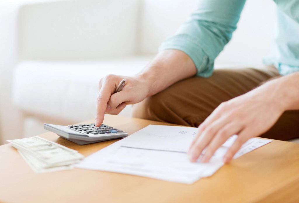 Closeup of man seated at coffee table, calculating bills using a handheld calculator, a small stack of cash, and a set of notes; concept photo for spousal maintenance calculations.