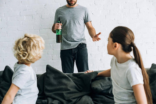 A man holding a bottle of alcohol and standing in front of two children, wondering, "How can mental health and substance abuse impact marriage and divorce?"